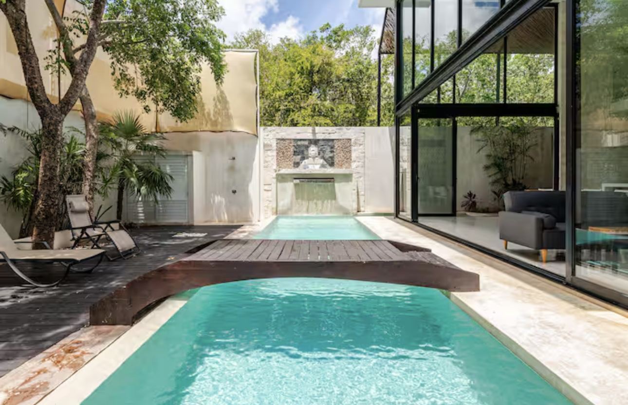 Pool with a wooden bridge and Buddha water feature in front of the glass Tulum villa